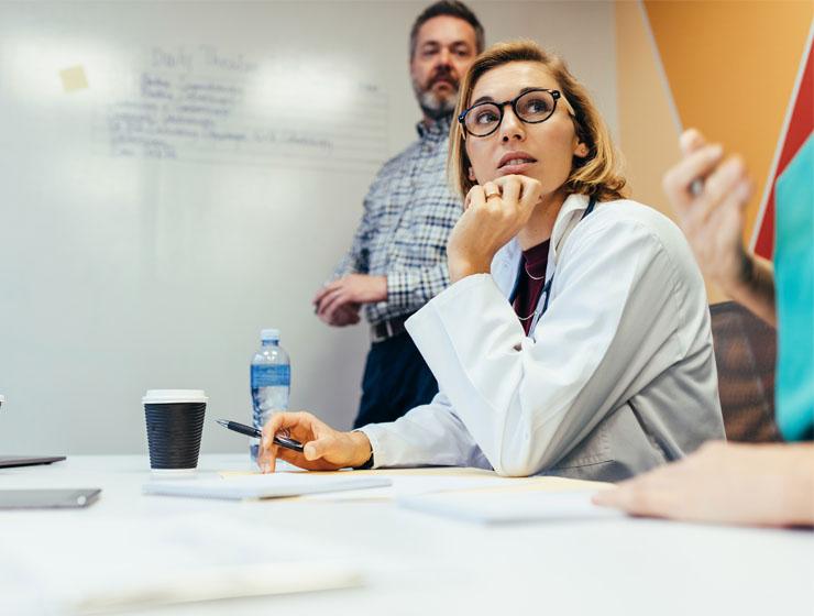 A female healthcare professional sits at a conference table with her chin resting in her hand, and elbow propped on the table. She is looking at someone speaking next to her. A male stands at a whiteboard behind her, looking at the speaker.