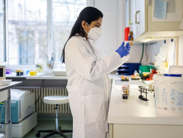 In a laboratory setting, a woman in a lab coat stands at a counter wearing gloves and a mask while holding a test tube.