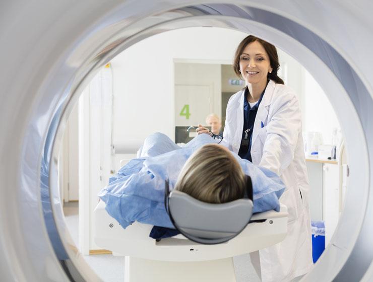 A female healthcare professional smiles while looking down at a patient who is laying down to get a scan. A male professional sits at a computer in the background.
